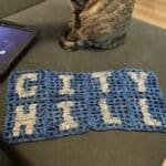 Blue crochet filet panels spelling “Hill City” laid out on a couch beside a tabby cat during the making process of a handmade sports cardigan by Pink Pearl Yarns.