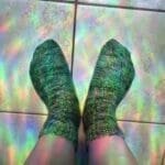 Hand-knit green socks worn on feet with rainbow sunlight reflections across the floor.